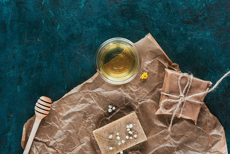 Wrapped cosmetics and soap sit on a blue table with a pot of honey.